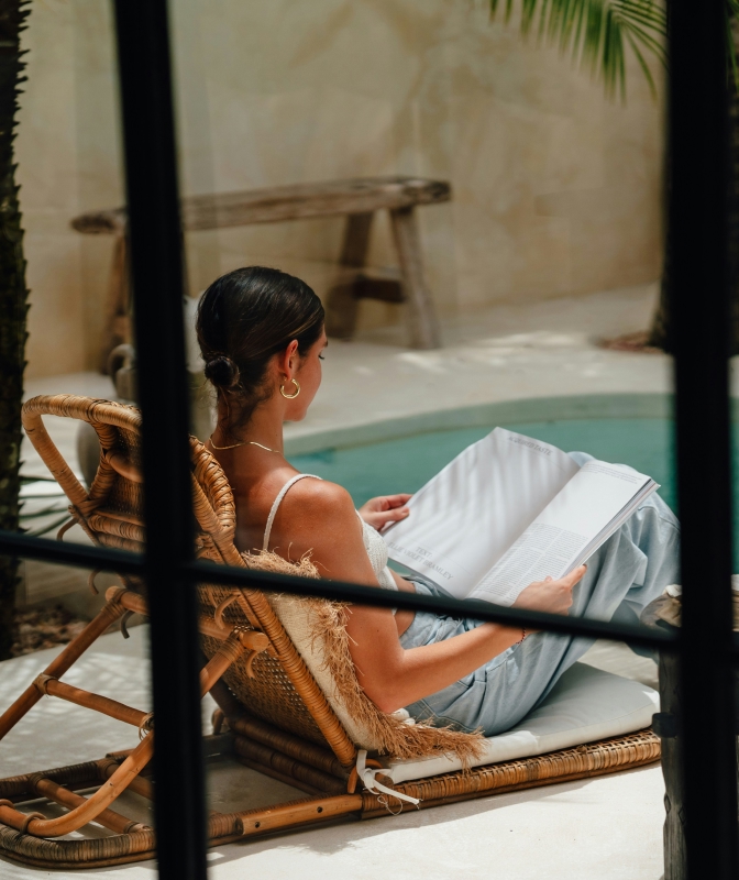 Woman relaxing in a wicker chair beside a pool, reading a book in a serene outdoor setting.